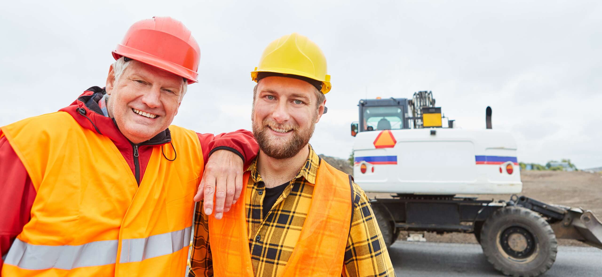 Older man resting his arm on the shoulder of a younger man, both in high visibility clothing with hard hats on. Construction machinery in the baackground